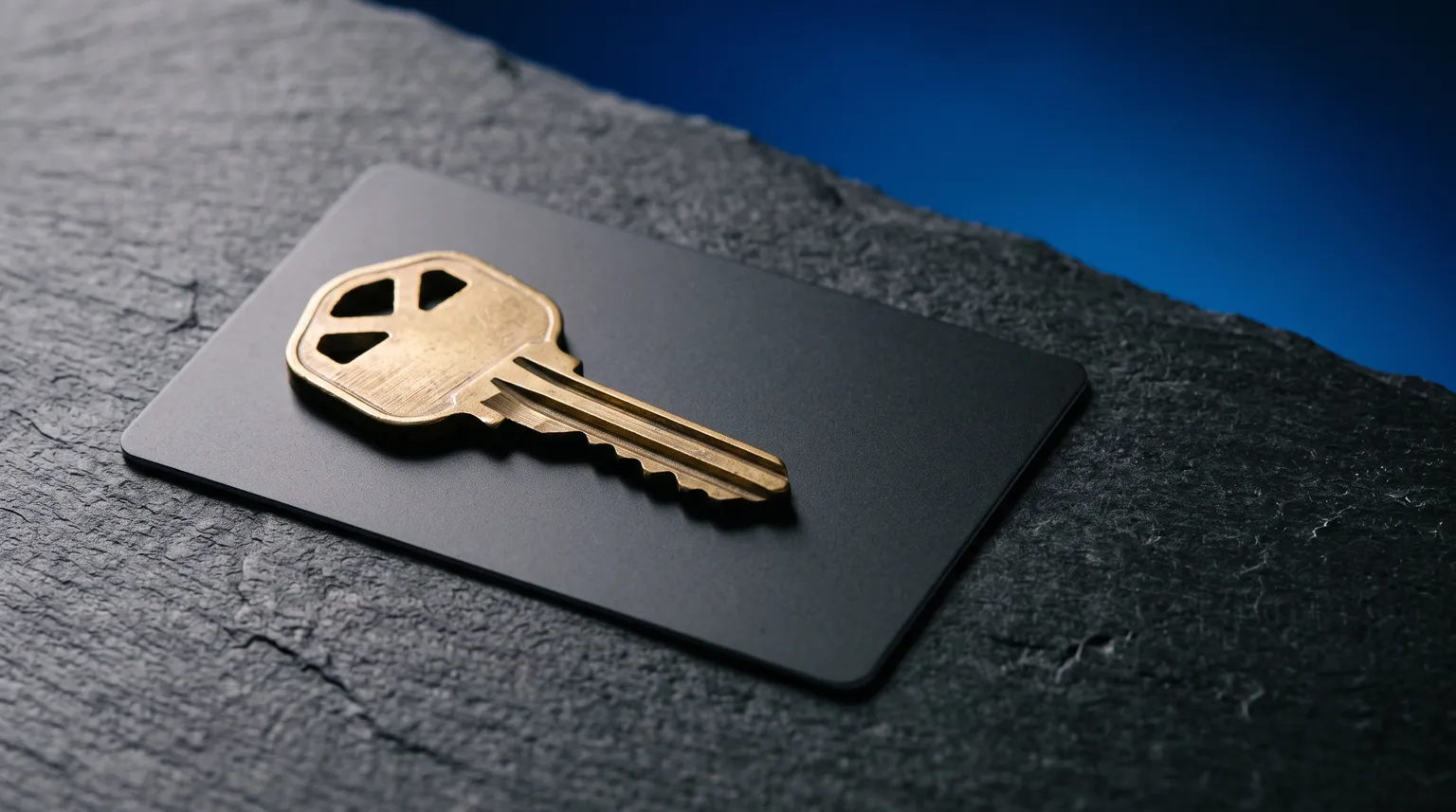 A brass house key resting on a plain dark credit card, lit against a deep blue background.