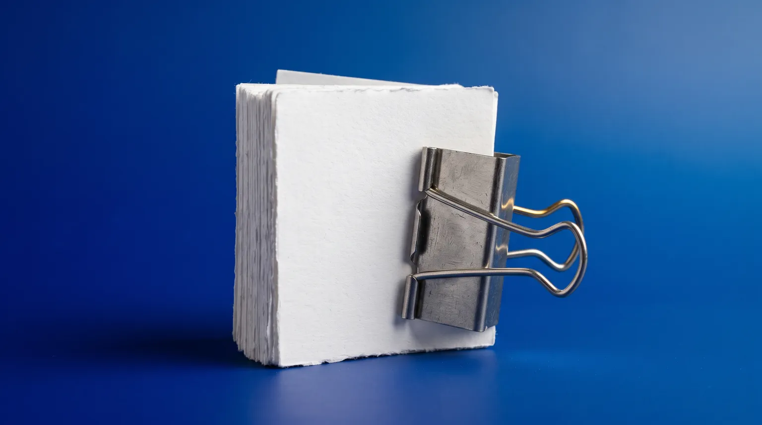 A thick stack of plain paper slips clamped in a heavy steel binder clip on a deep blue surface.